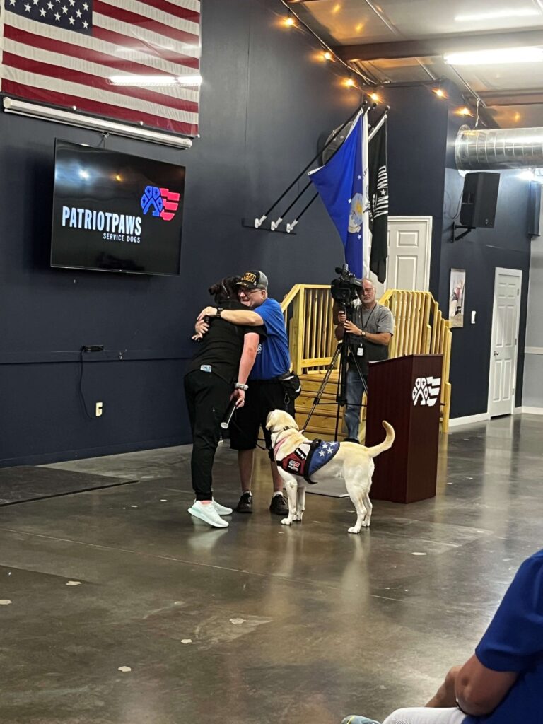 Two people hug at a Patriot PAWS event with a service dog beside them.